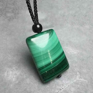 Puede incluir: Colgante rectangular de malaquita con una superficie verde pulida y rayada. El colgante está suspendido de un collar de cordón negro con un acento de cuentas negras. Los patrones naturales de la piedra crean un diseño único y llamativo.