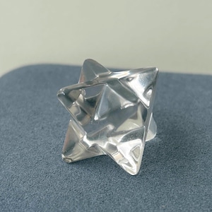 May include: A clear, faceted crystal star-shaped object. The object has a geometric design with multiple triangular faces, reflecting light. The star is set against a gray background, highlighting its shape and clarity.