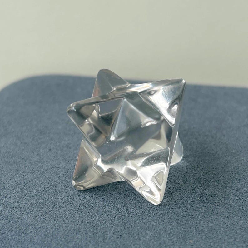 May include: A clear, faceted crystal star-shaped object. The object has a geometric design with multiple triangular faces, reflecting light. The star is set against a gray background, highlighting its shape and clarity.