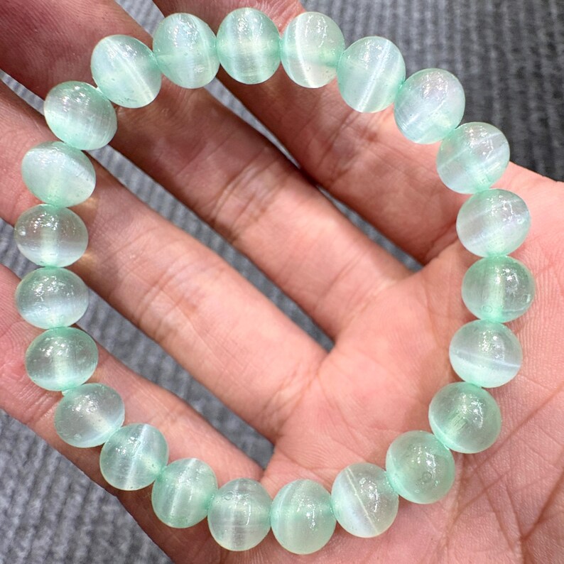 May include: A bracelet featuring round, light green beads with a translucent, striped appearance. The beads are strung together, forming a complete circle. The bracelet is shown on a person's hand.