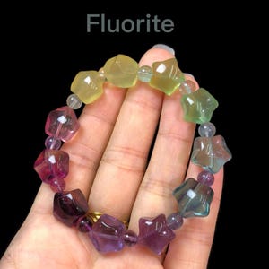 May include: A Fluorite bracelet with star-shaped beads in yellow, green, pink, and purple hues. The bracelet is strung with small, clear beads. The word "Fluorite" is displayed at the top. The bracelet is held against a black background.