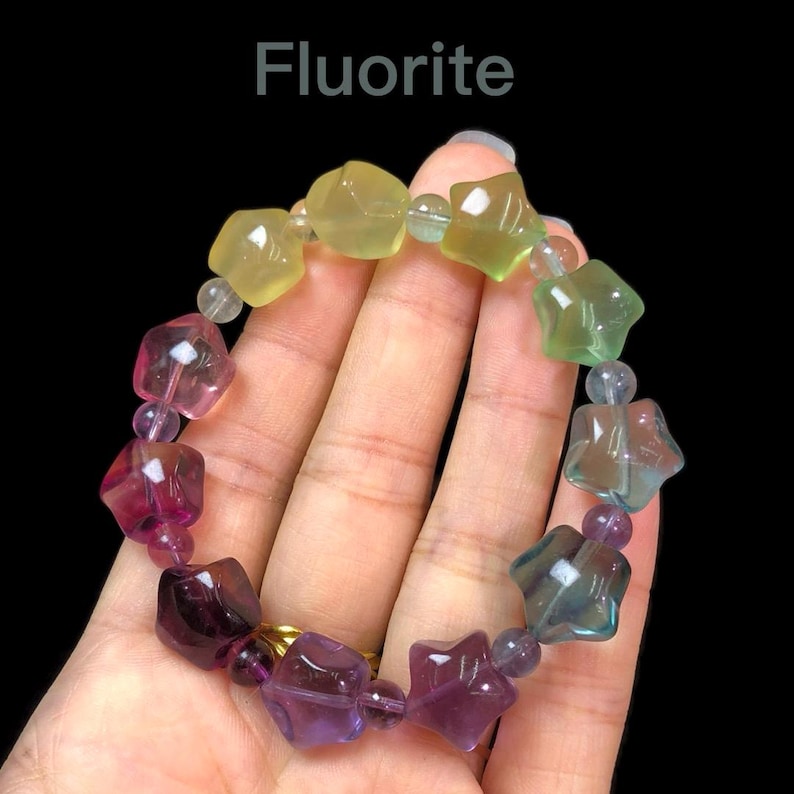 May include: A Fluorite bracelet featuring star-shaped beads in shades of yellow, green, pink, and purple. The bracelet is strung with small, clear beads and is held against a black background. The word "Fluorite" is displayed at the top.