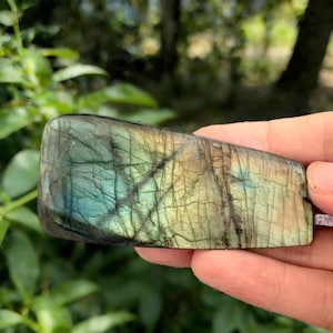 May include: A polished labradorite gemstone with a rectangular shape. The stone displays a mix of colors, including blues, greens, and yellows, with dark veining throughout. The gemstone is held in a hand against a blurred green background.