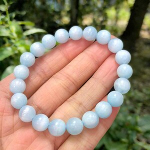 Op de afbeelding: Een armband van lichtblauwe aquamarijn edelstenen. De ronde kralen zijn aan elkaar geregen, waardoor een delicaat en elegant sieraad ontstaat. De armband wordt in een hand gehouden, waarbij de zachte, lichtblauwe kleur van de stenen wordt benadrukt.