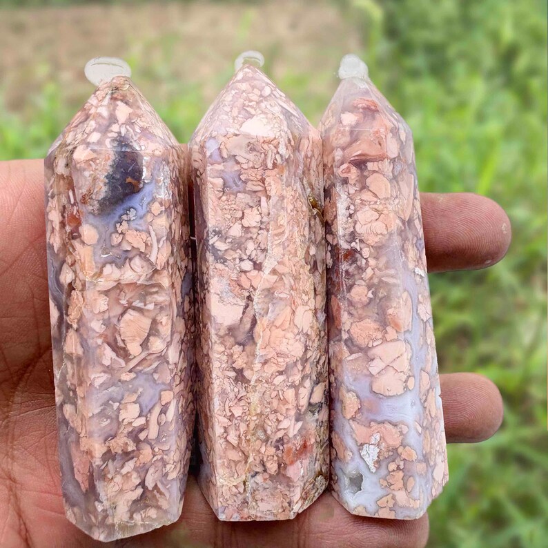 May include: Three natural stone obelisks with a mix of peach, cream, and gray tones. Each obelisk has a faceted shape and a clear crystal point at the top. The stones are held in a hand against a blurred green background.