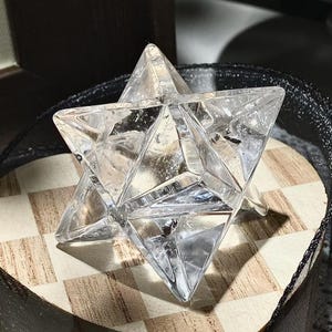May include: A clear quartz crystal Merkaba star. The geometric shape is composed of two intersecting tetrahedrons, creating a three-dimensional star. The crystal is displayed on a wooden surface with a black and white checkered pattern.