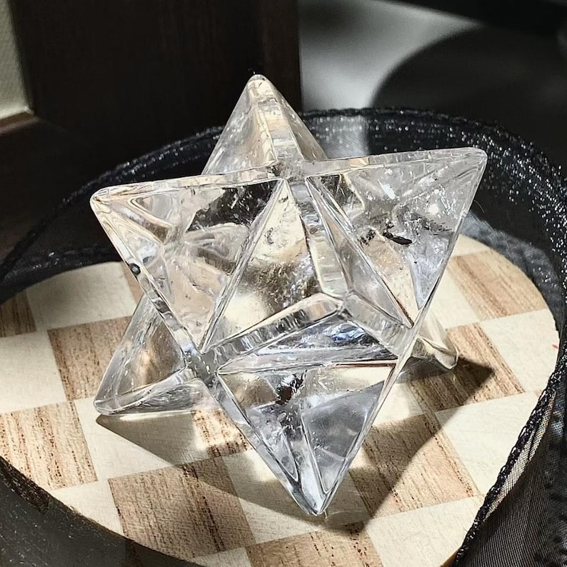 May include: A clear quartz crystal Merkaba star. The geometric shape is composed of two intersecting tetrahedrons, creating a three-dimensional star. The crystal is displayed on a wooden surface with a black and white checkered pattern.