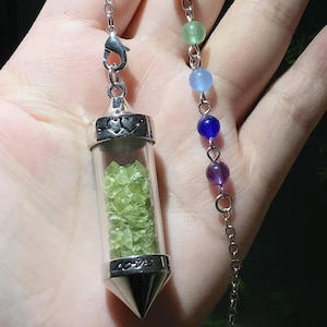 Puede incluir: Un colgante de cristal transparente lleno de pequeños cristales verde claro, suspendido de una cadena plateada. El colgante tiene una tapa plateada con diseños de corazón y una base puntiaguda. Una cadena separada presenta cuentas de colores.