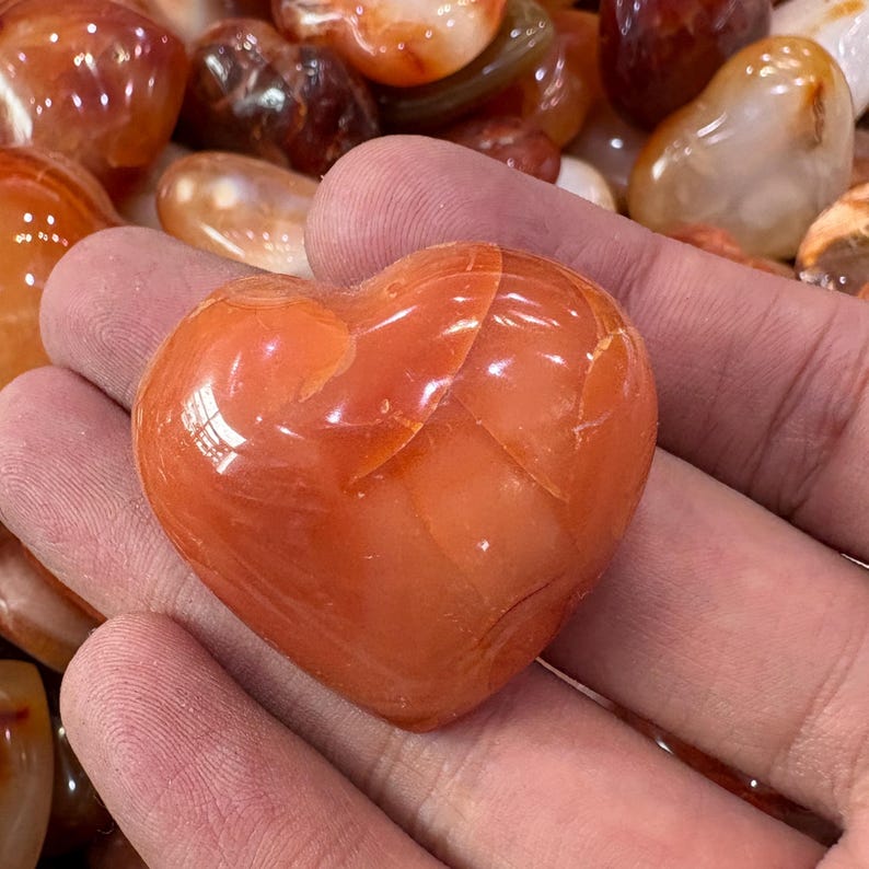 May include: A polished, heart-shaped carnelian stone, displaying a warm orange hue with natural veining. The stone is held in a hand, with other polished stones visible in the background. The heart-shaped stone is approximately 2 inches in length.