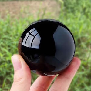 Pode incluir: Uma bola de cristal de obsidiana preta, polida e esférica. A superfície reflete a luz, criando reflexos brilhantes. A bola é segurada em uma mão contra um fundo verde desfocado de grama e folhagem.