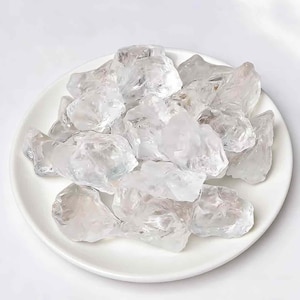 Puede incluir: Un plato de cerámica blanco lleno de cristales de cuarzo transparentes de forma irregular. Los cristales tienen un aspecto translúcido y varían en tamaño, creando una superficie texturizada. El plato es redondo y está sobre un fondo blanco.