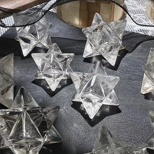 May include: Clear quartz Merkaba star crystals. These geometric, star-shaped crystals are faceted with multiple triangular planes. The crystals are displayed on a dark surface, with shadows adding depth. The Merkaba stars are a spiritual and metaphysical item.