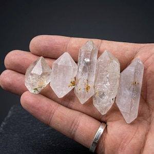May include: Five clear quartz crystal points of varying sizes are held in a hand. The crystals have natural inclusions and a polished appearance. A silver ring is visible on the hand. The background is dark.