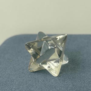 May include: A silver-colored, geometric star-shaped object with multiple triangular facets. The object is highly reflective, catching the light and creating highlights and shadows. The star is resting on a gray surface.