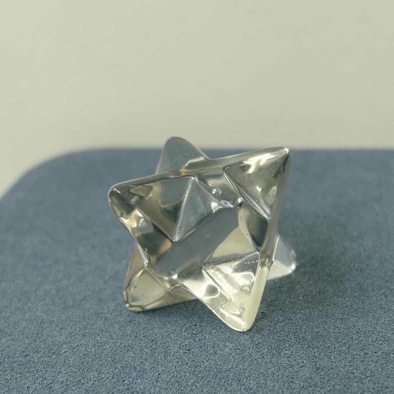May include: A silver-colored, geometric star-shaped object with multiple triangular facets. The object is highly reflective, catching the light and creating highlights and shadows. The star is resting on a gray surface.