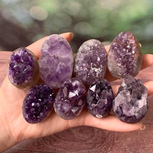 May include: A collection of eight small, oval-shaped amethyst crystal geodes with deep purple, faceted interiors and smooth, rounded outer shells. The crystals vary in shade from light lavender to dark violet, showcasing natural crystalline structures.