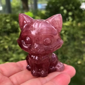 Könnte beinhalten: Eine geschnitzte, rot-violette Kristallkatzenfigur. Die Katze sitzt mit aufgestellten Ohren und weit geöffneten Augen. Die Figur ist glatt und poliert, mit sichtbaren Details im Stein.
