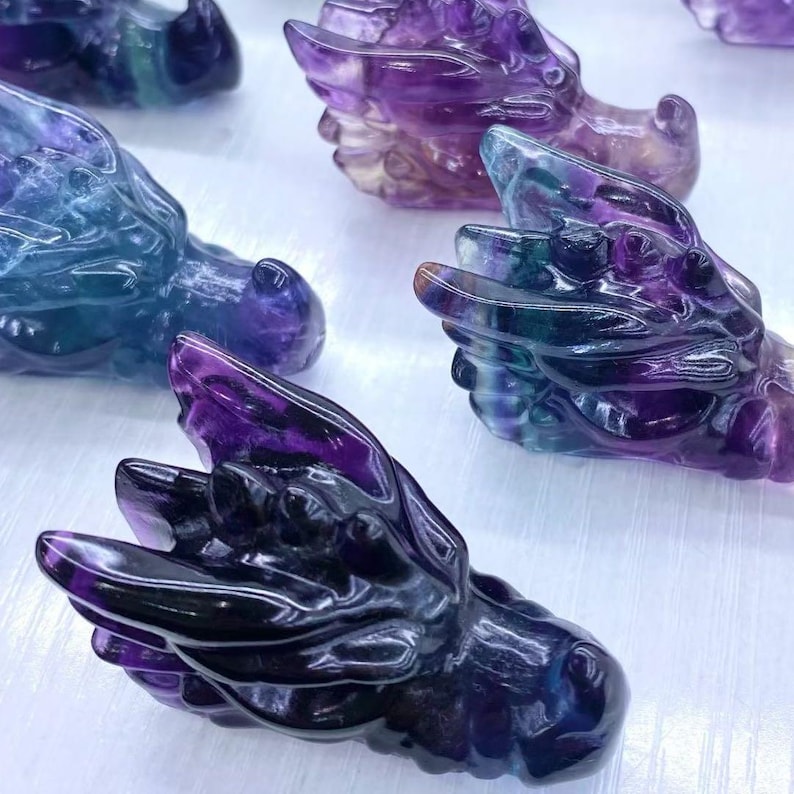 May include: Several carved dragon heads in shades of purple, blue, and green. The crystal sculptures feature detailed wings and a smooth, polished finish. The dragon heads are displayed on a light-coloured surface.