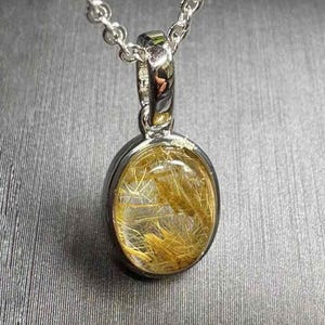 May include: A silver pendant necklace with an oval, clear gemstone containing golden, hair-like inclusions. The pendant is set in a silver bezel and suspended from a delicate silver chain. The gemstone has a polished, reflective surface.