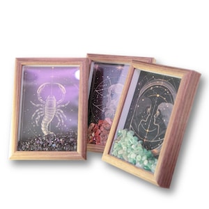 May include: Three framed art pieces with wooden frames. Each frame contains a different zodiac-themed illustration with a background of stars and a pile of colorful stones at the bottom. The art pieces are angled towards the viewer.