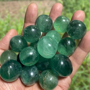Può includere: Una collezione di sfere di fluorite verde lucidata. Le sfere variano in tonalità di verde, da chiaro a scuro, e hanno una superficie liscia e lucida. Le sfere sono tenute in una mano, con uno sfondo sfocato di fogliame verde.