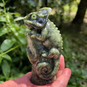 Könnte beinhalten: Eine detaillierte Skulptur eines Chamäleons in Grün- und Schwarztönen, das auf einem Baumstamm sitzt. Das Chamäleon hat einen strukturierten Körper, einen eingerollten Schwanz und ein detailliertes Gesicht mit einem markanten Auge. Die Skulptur wird in einer Hand gehalten.