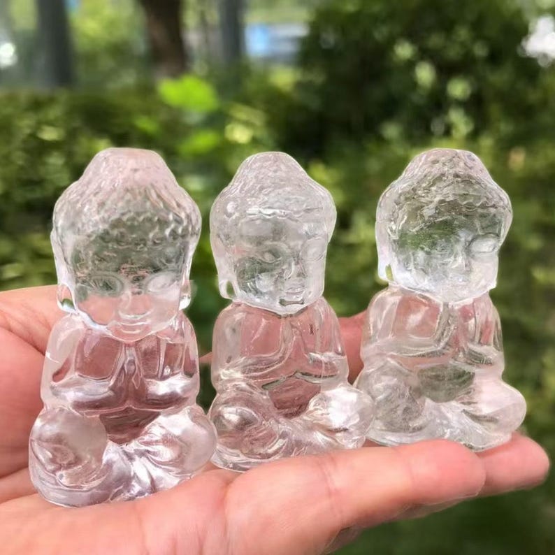 May include: Three clear quartz Buddha figurines in a row, each in a seated meditation pose. The statues are held in a hand, with a blurred green background.