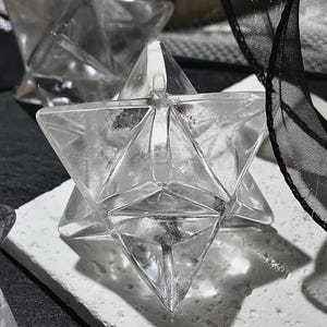 May include: Clear crystal Merkaba star. The geometric shape is composed of two intersecting tetrahedrons, creating a three-dimensional star. The crystal is placed on a white surface, with a black ribbon in the background.