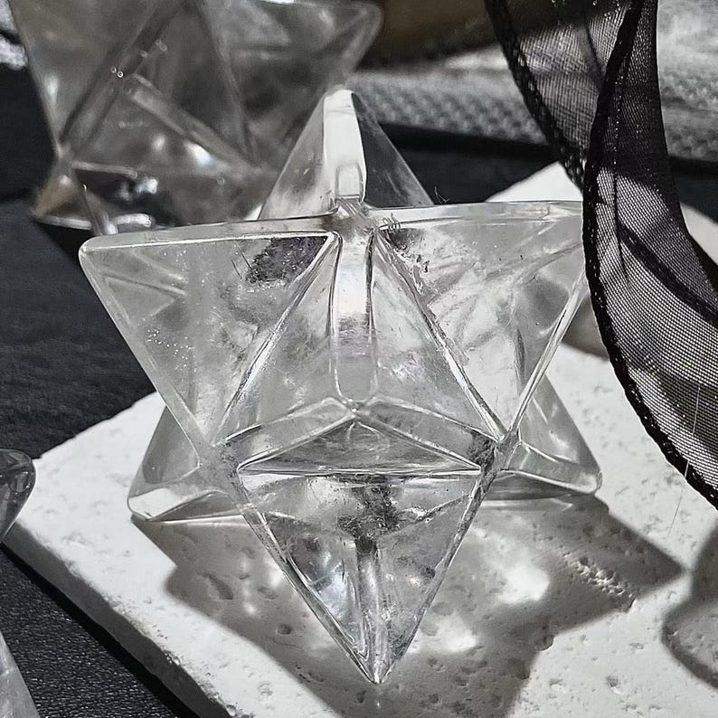 May include: Clear crystal Merkaba star. The geometric shape is composed of two intersecting tetrahedrons, creating a three-dimensional star. The crystal is placed on a white surface, with a black ribbon in the background.