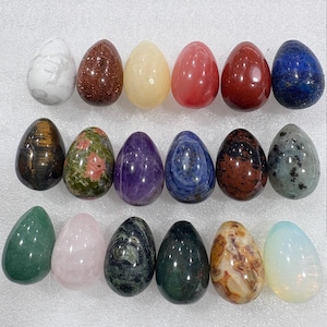 May include: A collection of sixteen polished gemstone eggs in various colors and patterns. The eggs include white marble, goldstone, rose quartz, amethyst, and other stones. Each egg is approximately 2 inches tall.