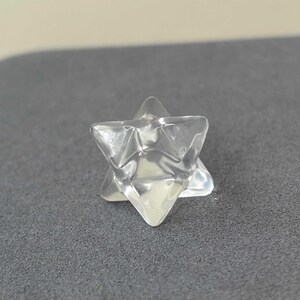 May include: A clear, faceted crystal Merkaba star. The star is transparent and reflects light, with a six-pointed star shape. The crystal is set against a gray background.