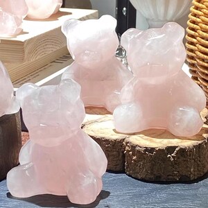 May include: Several rose quartz bear figurines. The bears are carved from translucent pink stone and sit on small wooden slices. The carvings have smooth, rounded features, and the light reflects off their polished surfaces.