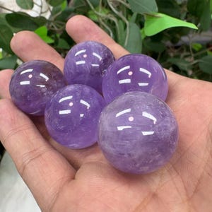 May include: Five polished amethyst spheres, each displaying a rich purple hue with subtle variations in tone. The spheres are held in an open hand, showcasing their smooth, rounded forms and natural crystalline patterns. The spheres are a popular item in the crystal and gemstone category.