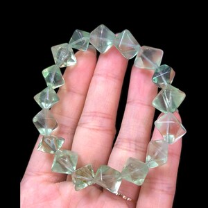 May include: A light green fluorite bracelet. The bracelet is composed of faceted, square-shaped beads. The beads are translucent, with visible internal structures. The bracelet is held against a dark background, highlighting the color and clarity of the stones.