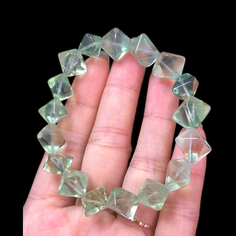May include: A light green fluorite bracelet. The bracelet is composed of faceted, square-shaped beads. The beads are translucent, with visible internal structures. The bracelet is held against a dark background, highlighting the color and clarity of the stones.