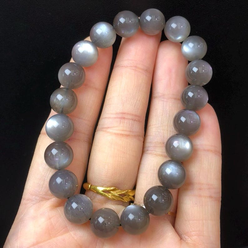 May include: A bracelet made of grey moonstone beads. The bracelet is composed of spherical, polished beads in different shades of grey, with a soft lustre. The beads are strung together, forming a complete circle. The bracelet is held against a dark background.