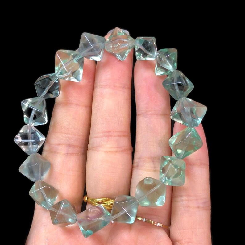 May include: A bracelet featuring translucent, square-cut beads in shades of light blue and green. The beads are strung together, forming a circular shape. The bracelet is held against a dark background.