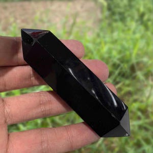 Pode incluir: Uma ponta de cristal de obsidiana preta polida, segurada em uma mão. O cristal tem uma forma retangular com extremidades pontiagudas. A superfície reflete a luz, mostrando seu acabamento brilhante. O fundo é folhagem verde desfocada.