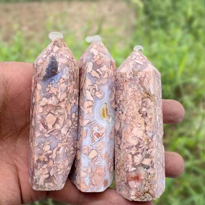 May include: Three polished stone obelisks with a light pink and beige mottled pattern. Each has a clear, rounded top. The stones are held in a hand against a blurred green background.