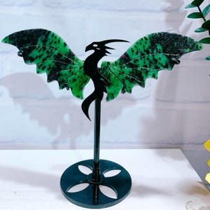 May include: A dragon ornament featuring a black silhouette body and green wings with black markings. The dragon is mounted on a black metal stand with a circular base. The sculpture is displayed on a white surface.