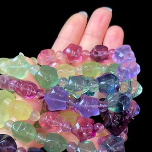 May include: A collection of colorful gemstone bracelets. The beads are various shapes and sizes, including star and irregular shapes, in shades of pink, purple, green, yellow, and blue. The bracelets are strung on a clear elastic cord.