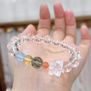 May include: Clear quartz crystal bracelet with a mix of colorful gemstone beads. The bracelet features a star-shaped crystal charm and a variety of round beads in shades of blue, yellow, orange, and a speckled brown. The bracelet is held in a hand.