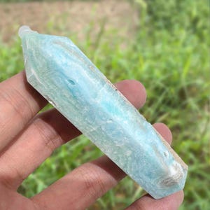May include: A light blue crystal point with a hexagonal shape. The crystal has a textured surface with variations in color, ranging from pale to medium blue. The crystal is held in a hand against a blurred green background.