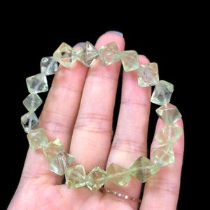 May include: A light green gemstone bracelet featuring square-cut stones. The bracelet is strung together, creating a circular shape. The stones have a translucent quality, and the bracelet is displayed against a black background.