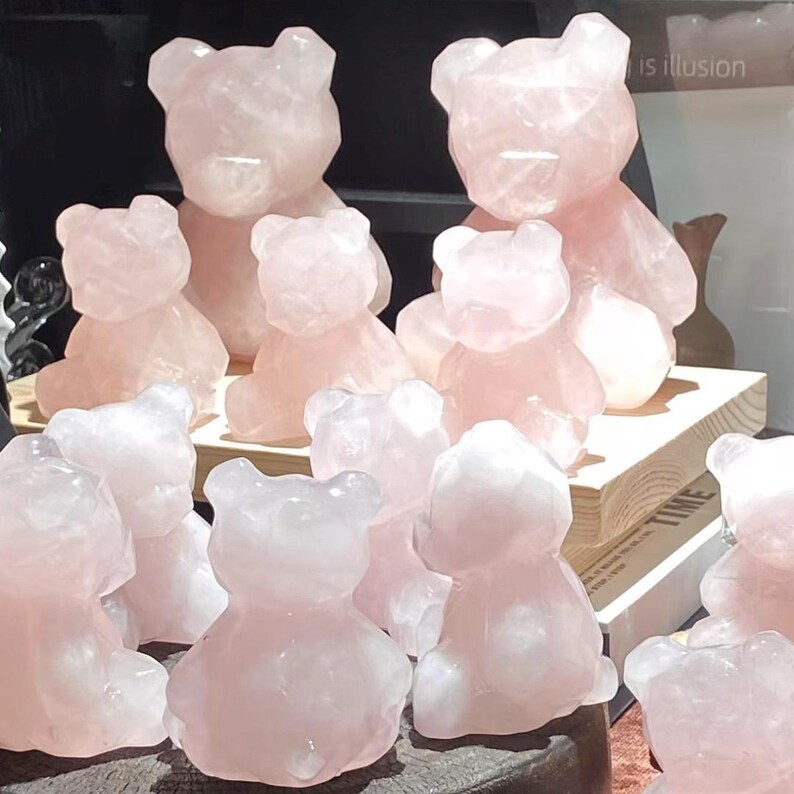 May include: A collection of rose quartz bear figurines in various sizes. The bears are a soft pink color and have a faceted, crystalline appearance. Some bears are sitting, while others are standing. The background is dark, with a wooden shelf.