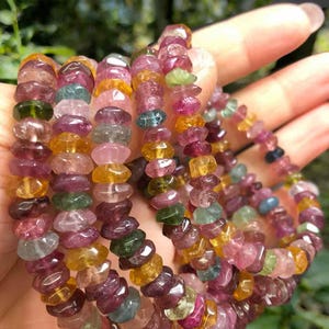 May include: A collection of colorful tourmaline gemstone bracelets. The beads are a mix of pink, green, yellow, and purple. The bracelets are strung together, showcasing the natural variations in color and shape of the stones.