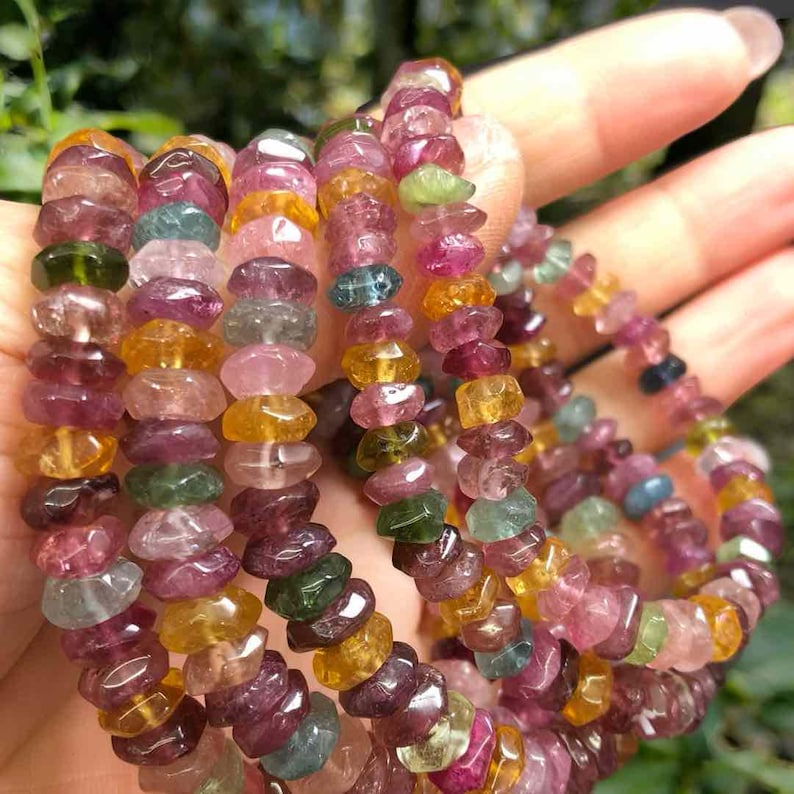 May include: A collection of colorful tourmaline gemstone bracelets. The beads are a mix of pink, green, yellow, and purple. The bracelets are strung together, showcasing the natural variations in color and shape of the stones.