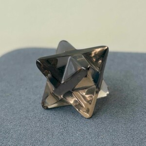 May include: A faceted, star-shaped object with a metallic, reflective surface. The object has a geometric design with multiple triangular faces. The color appears to be a mix of silver and dark gray, with light reflecting off the surfaces.