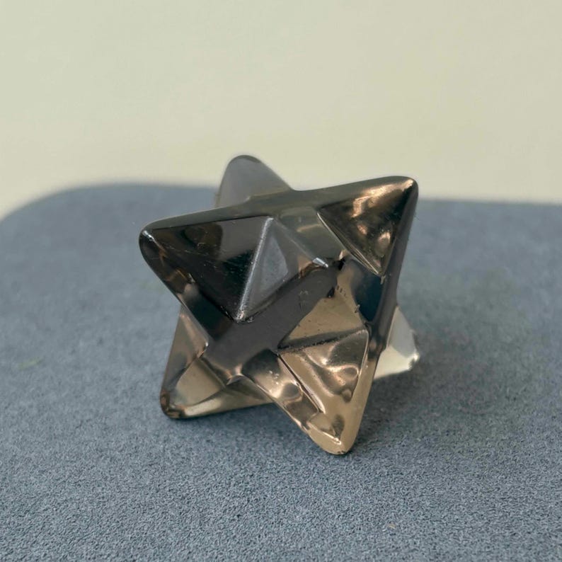 May include: A faceted, star-shaped object with a metallic, reflective surface. The object has a geometric design with multiple triangular faces. The color appears to be a mix of silver and dark gray, with light reflecting off the surfaces.