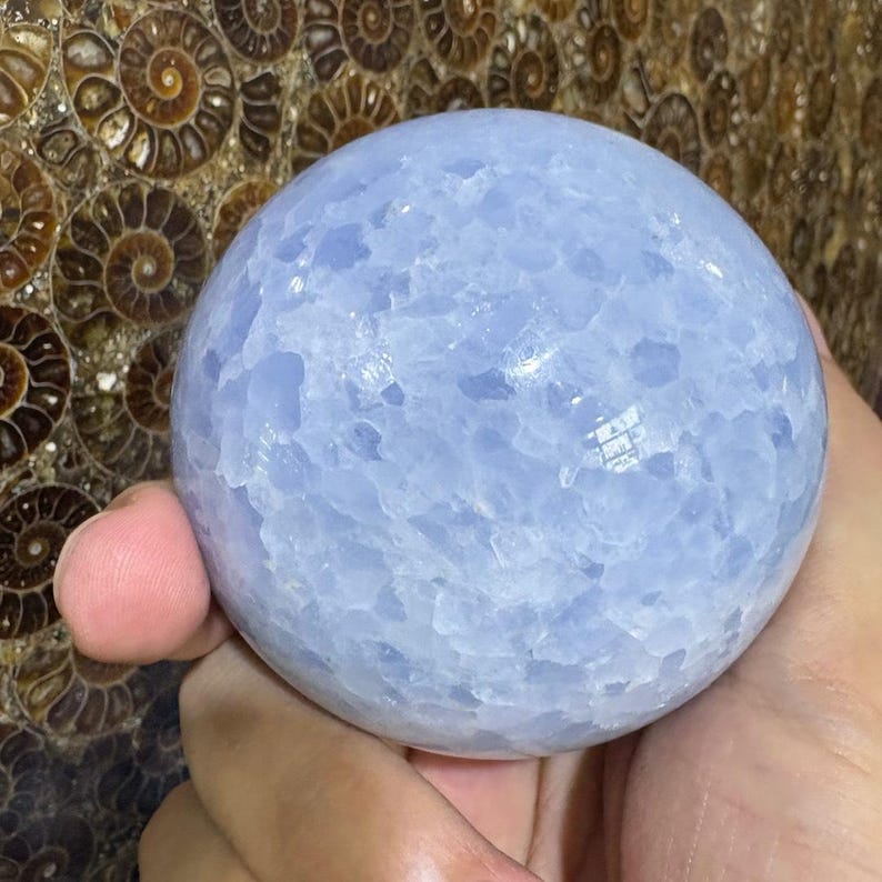 May include: A light blue sphere with a textured surface. The sphere is held in a hand, with a background of brown and tan spiral patterns. The sphere appears to be made of a crystalline material.